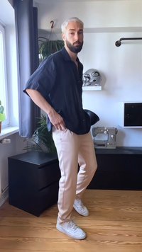Homem com cabelo e barba descoloridos, vestindo uma camisa escura, calças claras e ténis brancos, de pé dentro de casa perto de uma janela e móveis pretos.
