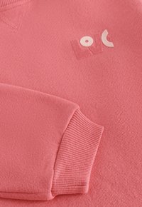 Różowy sweter z miękką fakturą, ściągaczami przy rękawach i subtelnym białym logo z zaokrąglonymi literami na klatce piersiowej.