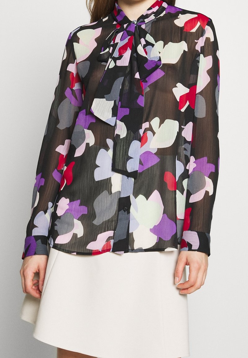 Femme portant un chemisier noir transparent à manches longues avec des imprimés floraux abstraits en violet, rouge, blanc et gris, associé à une jupe blanche.