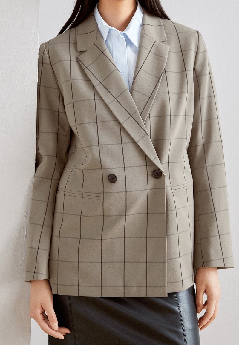 Femme portant un blazer beige à double boutonnage à carreaux, une chemise bleu clair et une jupe en cuir noire, debout contre un mur blanc.