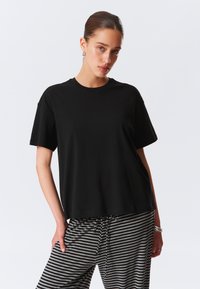 Juodas trumpomis rankovėmis marškinėliai iš minkštos medžiagos, pasižymintys laisvu siluetu ir apvaliu iškirpimu, derinami su pilkais ir juodais dryžuotais kelnėmis.