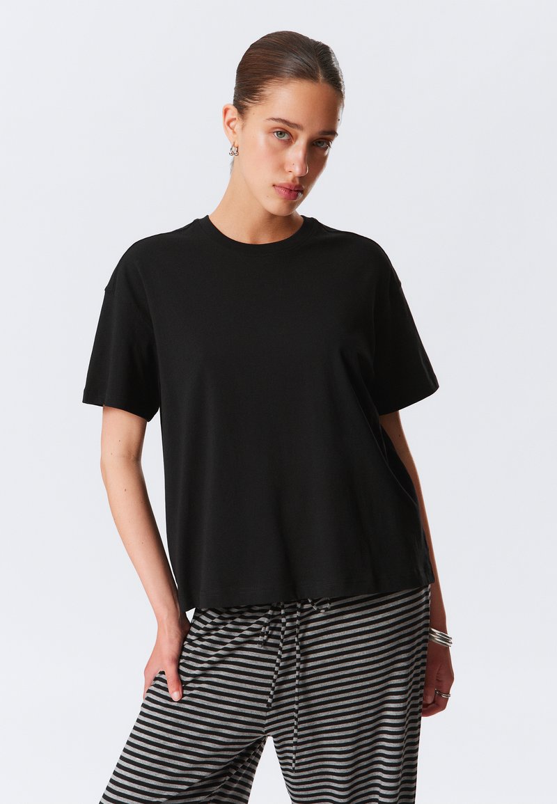 Juodas trumpomis rankovėmis marškinėliai iš minkštos medžiagos, pasižymintys laisvu siluetu ir apvaliu iškirpimu, derinami su pilkais ir juodais dryžuotais kelnėmis.