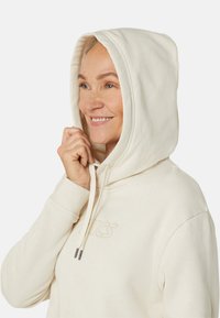 Cream-colored hoodie with a fleece texture, a drawstring hood, and embroidered logo. Features a relaxed fit and ribbed cuffs.