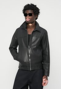 Black leather jacket with a glossy finish, zip closure, and a folded collar. Worn with dark clothing and sunglasses.