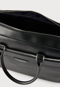 Sac à main Lacoste en cuir noir texturé avec fermetures éclair argentées et poignées arrondies, présenté partiellement ouvert sur un fond blanc.