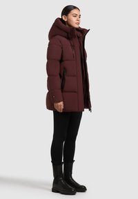 Burgundi puffertakaró magas gallérral, cipzáros elülső résszel, oldalsó zsebekkel és texturált, steppelt mintázattal. Fekete leggingsszel és csizmával párosítva.