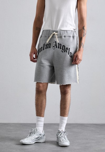 Osoba ubrana w szare szorty Palm Angels, białe skarpetki, szare sneakersy i białą koszulkę bez rękawów, stojąca na szarym podłodze w tle szarej ściany.