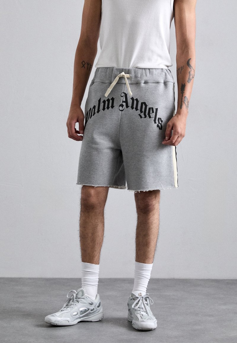 Person wearing gray Palm Angels shorts, white socks, gray sneakers, and a white sleeveless shirt, standing on gray floor against a gray wall.