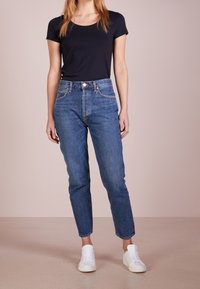 Modré denimové džíny s vysokým pasem a uvolněným střihem, kombinované s černým tričkem s krátkým rukávem a bílými teniskami na neutrálním pozadí.