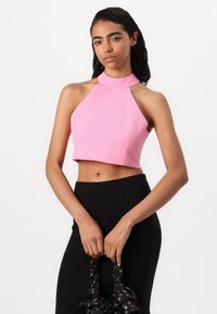 Roza halter crop top z oblikovanim silhueto, teksturirano tkanino in visokim ovratnikom. V kombinaciji z črnimi visokimi hlačami z rebrasto strukturo in cvetlično torbico.