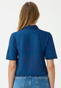 Frau mit langen blonden Haaren, die ein strukturiertes, kurzärmliges blaues Strickoberteil und ausgebleichte, hochtaillierte blaue Jeans trägt, von hinten gesehen.