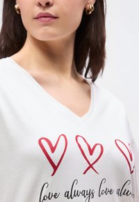 Γυναίκα που φοράει λευκό μπλουζάκι V λαιμόκοψη με τρεις κόκκινες καρδιές και μερικό καλλιγραφικό κείμενο που γράφει "πάντα αγάπη", χρυσά σκουλαρίκια κρίκοι, καστανά μαλλιά.