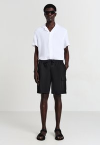 Camicia bianca a maniche corte con bottoni, pantaloni cargo neri con elastico in vita e tasche laterali, e sandali neri. Sfondo neutro.