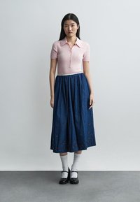 Mladá žena stojí oblečená v krátkym ružovom tričku s gombíkmi, modrej midi sukni s kvetinovými výrezmi, bielych ponožkách a čiernych topánkach Mary Jane.