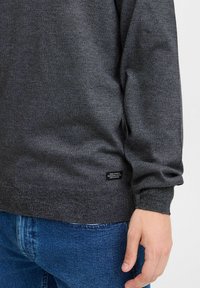 Close-up van een persoon in een donkergrijze gestructureerde trui en blauwe jeans, met een klein zwart label op de onderrand van de trui.