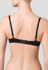 Bagfra af en person iført en sort bh med justerbare stropper og tre-huls lukning mod en ensfarvet baggrund.