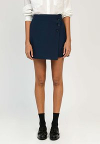 Jupe portefeuille bleu marine avec trois accents de boutons proéminents, forme ajustée, texture lisse, portée avec des chaussettes noires et des mocassins brillants.