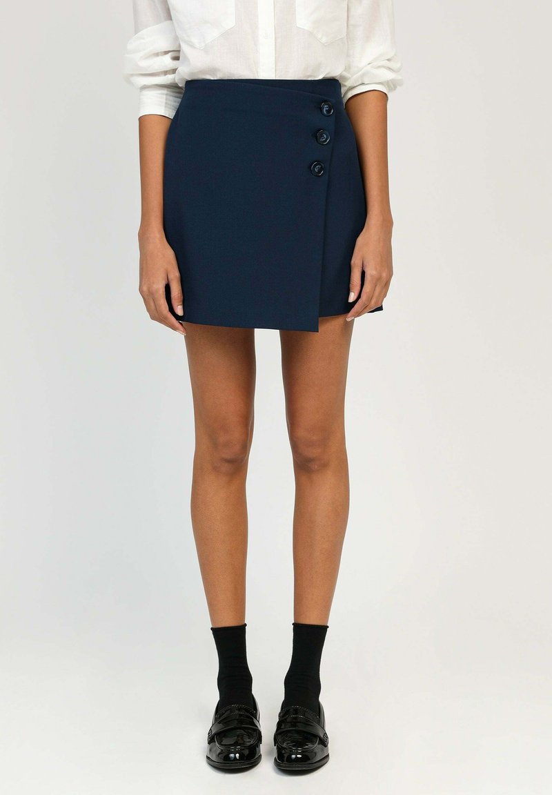 Jupe portefeuille bleu marine avec trois accents de boutons proéminents, forme ajustée, texture lisse, portée avec des chaussettes noires et des mocassins brillants.