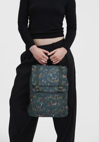 Zeleni ruksak s uzorkom koji prikazuje životinje i lišće, s prednjim džepom na zatvarač i podesivim trakama. Izrađen od izdržljivog materijala.