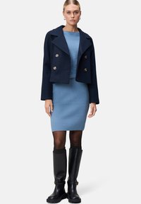 Marineblauwe, korte, dubbelzijdige blazer over een lichtblauwe geribde jurk, gecombineerd met kniehoge zwarte laarzen en zwarte sokken. Stevige texturen.
