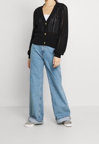 Cardigan nero lavorato a maglia con scollo a V, chiusura con bottoni e maniche lunghe. Abbinato a jeans blu a vita alta e dalla vestibilità ampia, e scarpe bianche e nere.