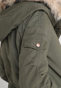 Olivově zelená parka s umělou kožešinou na límci, obsahující kapsu na rukáv se zapínáním na knoflíky a zpevněné švy, navržená pro odolnost a teplo.