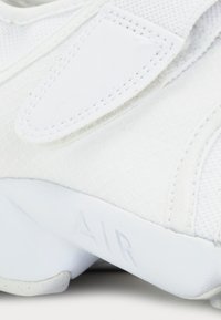White athletic shoe featuring a textured mesh upper, smooth leather accents, and a raised "AIR" detail on the sole. Velcro strap closure.