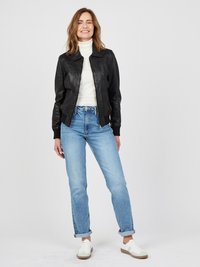 Schwarze Lederbomberjacke, weißer Rollkragenpullover, hellblaue Jeans und weiße Sneakers. Gerade Passform mit umgeschlagenen Bündchen und minimalem Hardware.
