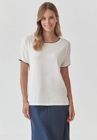 White short-sleeve top with a round neckline and black trim. Smooth fabric, relaxed fit, paired with a dark blue skirt.