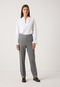 Vrouw staat tegen een effen achtergrond in een witte blouse met lange mouwen, grijze pantalon en bruine loafers, recht vooruit kijkend met een ontspannen houding.