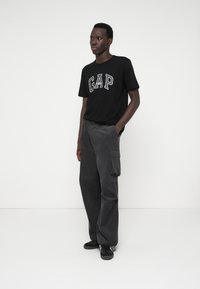 Czarny bawełniany t-shirt z szarym logo "GAP", zestawiony z szarymi spodniami cargo z korduroju z bocznymi kieszeniami. Model stoi na tle jednolitym.