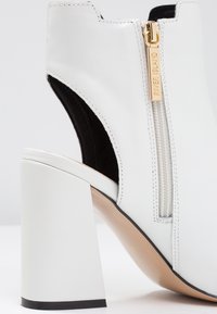 Botín de tacón de cuero blanco con cremallera lateral y tirador dorado. Presenta un tacón cuadrado y grueso, además de un diseño con parte trasera abierta.
