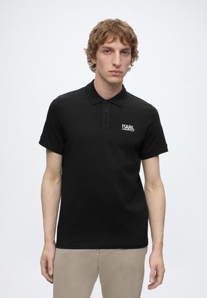 Młody mężczyzna o kręconych jasnobrązowych włosach, ubrany w czarną koszulkę polo z krótkim rękawem i beżowe spodnie na jednolitym białym tle.