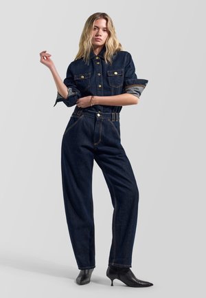 Donna bionda che indossa una tuta in denim scuro con bottoni, maniche arrotolate e stivali neri con tacco a punta, in posa davanti a uno sfondo semplice.