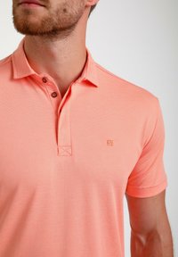 Kortärmad polotröja i ljus korallfärg, tillverkad av textil med struktur. Har en knappslå och en liten logotyp på bröstet.