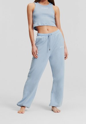 Vrouw draagt een lichtblauw mouwloos crop top en bijpassende losse joggingbroek, staat blootsvoets op een effen achtergrond.
