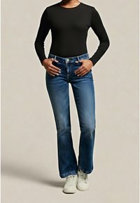 Personne portant un haut noir à manches longues, un jean bootcut bleu et des baskets blanches, debout les mains dans les poches.