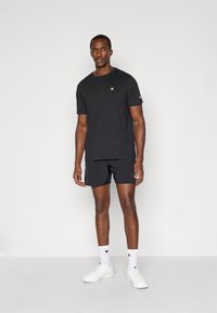 Camiseta negra de manga corta con un logo en el lado izquierdo del pecho, combinada con pantalones cortos negros y zapatillas blancas, de pie contra un fondo neutro.