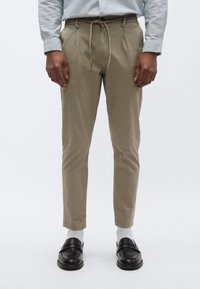 Χακί chinos με ελαστική μέση και κορδόνι, στενή εφαρμογή και πτυχώσεις μπροστά. Φοριούνται με μαύρα loafers και λευκές κάλτσες.