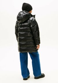 Doudoune noire avec finition brillante, capuche et design matelassé horizontal. Portée avec un jean bleu décontracté et des bottes noires, vue de dos.