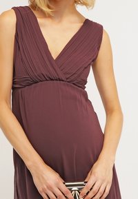 V-ringad, ärmlös klänning i djup plommonlila med plisserad byst och empiremidja. Modellen håller en liten, metallisk clutchtaske med geometrisk design.