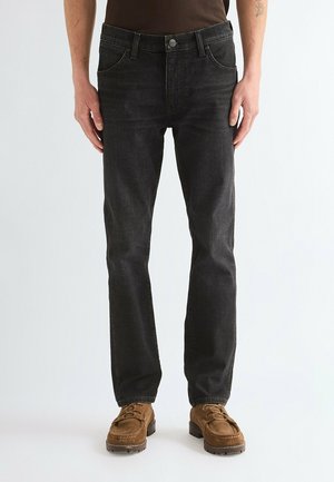 Man wearing black straight-leg jeans and brown suede lace-up shoes standing against a plain light background.