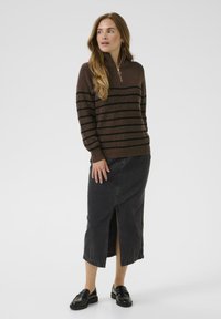 Brauner gerippter Pullover mit schwarzen Streifen und Reißverschlusskragen, kombiniert mit einem schwarzen Denimrock mit Schlitz vorne. Getragen mit schwarzen Slippern.