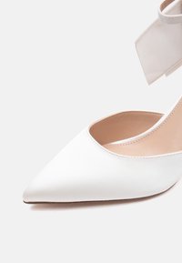 Chaussure à talon haut blanche à bout pointu avec bride cheville et détail nœud sur fond blanc uni.