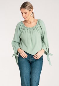 Jasnozielona, długo rękawowa bluzka z elastycznym dekoltem i detalami w postaci wiązań przy rękawach, w połączeniu z dżinsami z wysokim stanem w kolorze niebieskim. Miękka faktura.