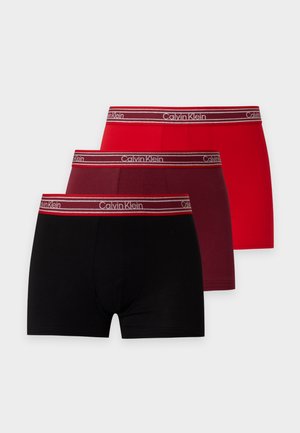 Jeune homme debout pieds nus, portant un boxer orange avec une ceinture de marque, sur un fond clair uni.