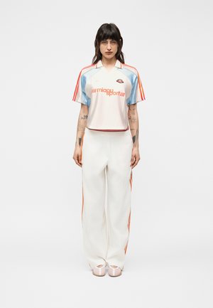 Femeie stând cu fața înainte, purtând un tricou sport alb cu accente albastre și portocalii și pantaloni albi cu dungi laterale portocalii.