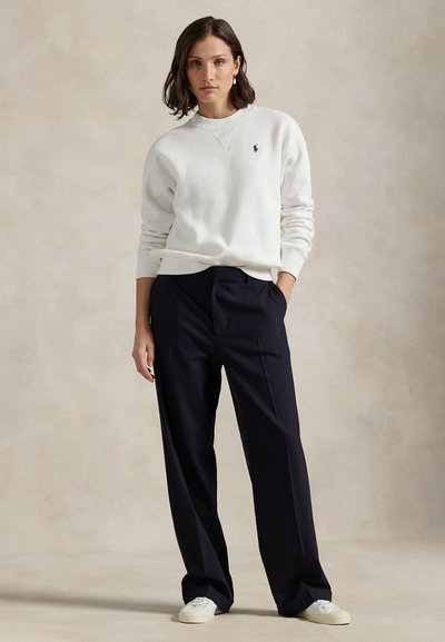 Γυναίκα που στέκεται με το χέρι στην τσέπη, φορώντας λευκό φούτερ, μαύρο παντελόνι με φαρδιά πόδια, λευκά αθλητικά παπούτσια και σκουλαρίκια πέρλες.