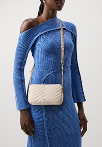 Beige quiltet.crossbody-veske med chevron-mønster, gullkjede-rem og glatt lærfinish, båret over en blå strukturert off-shoulder-kjole.