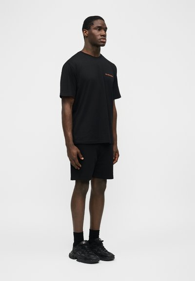 Ανδρας που στέκεται φορώντας μαύρο μπλουζάκι με μικρό πορτοκαλί κείμενο, μαύρη βερμούδα, μαύρες κάλτσες και μαύρα αθλητικά παπούτσια, μπροστά σε απλό γκρι φόντο.